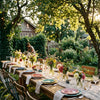 Een prachtig gedekte tuintafel met kleurrijk servies, verse bloemen en sfeervolle lichtslingers, badend in warm gouden uur zonlicht, klaar voor gasten.
